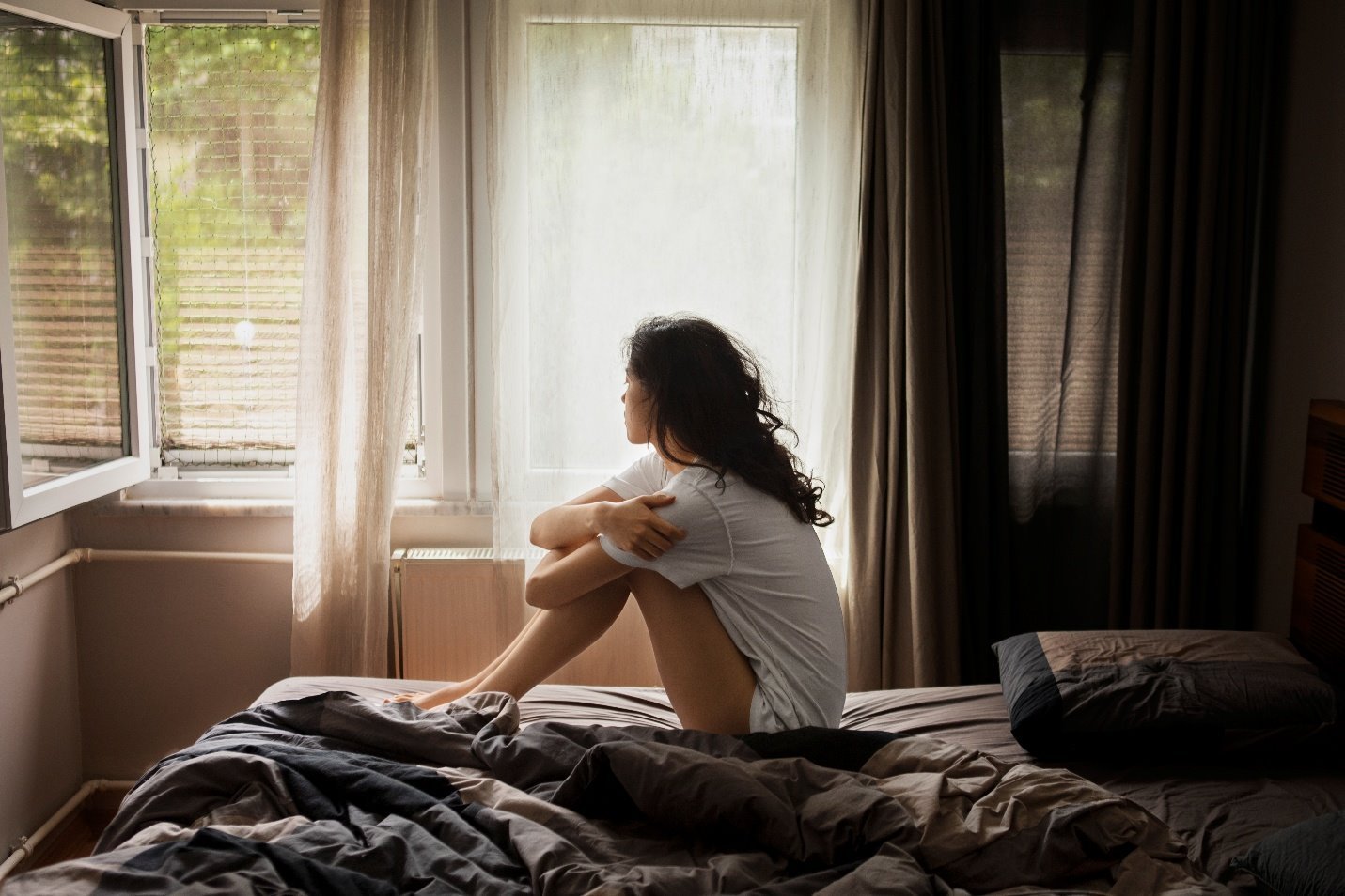 A woman sitting on a bed, looking out a bright window